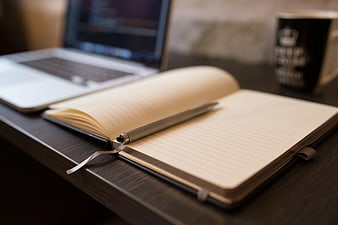 Open notebook with pen on desk beside laptop and coffee