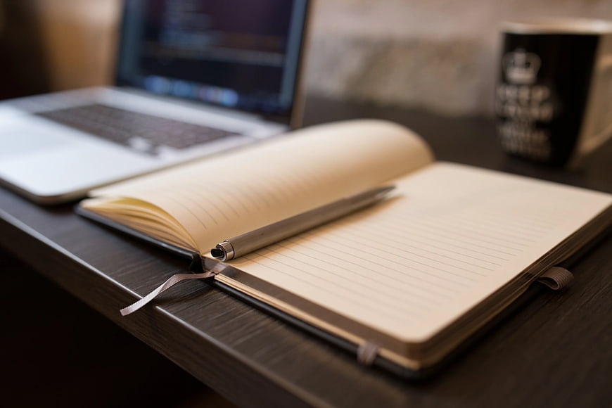 Open notebook with pen on desk beside laptop and coffee