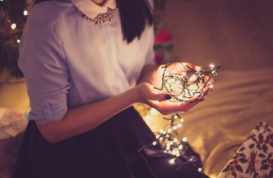 Woman holding glowing Christmas lights in cozy bedroom