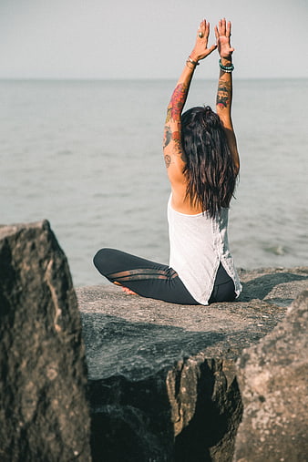 Woman with tattoos meditating on rocks by the ocean