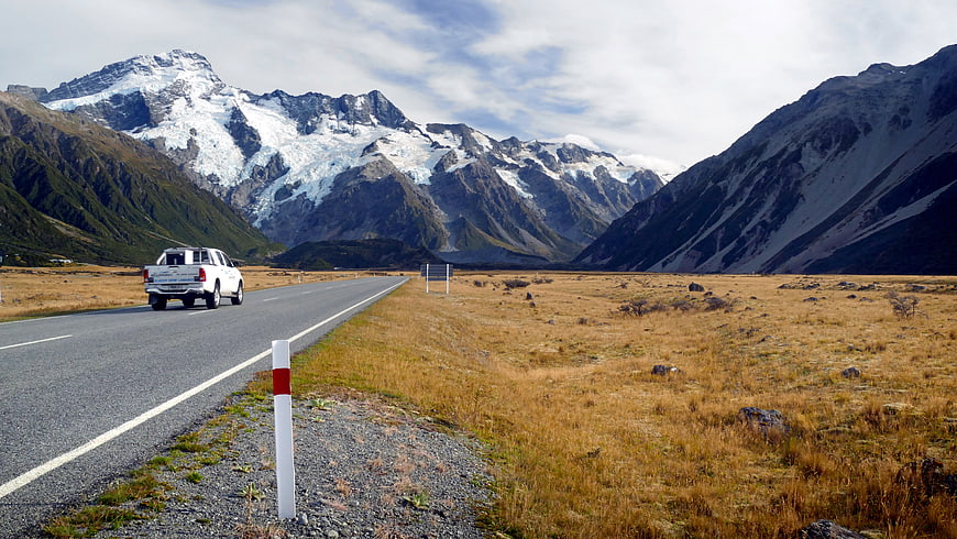 White pickup truck driving on scenic mountain road with snow-capped peaks