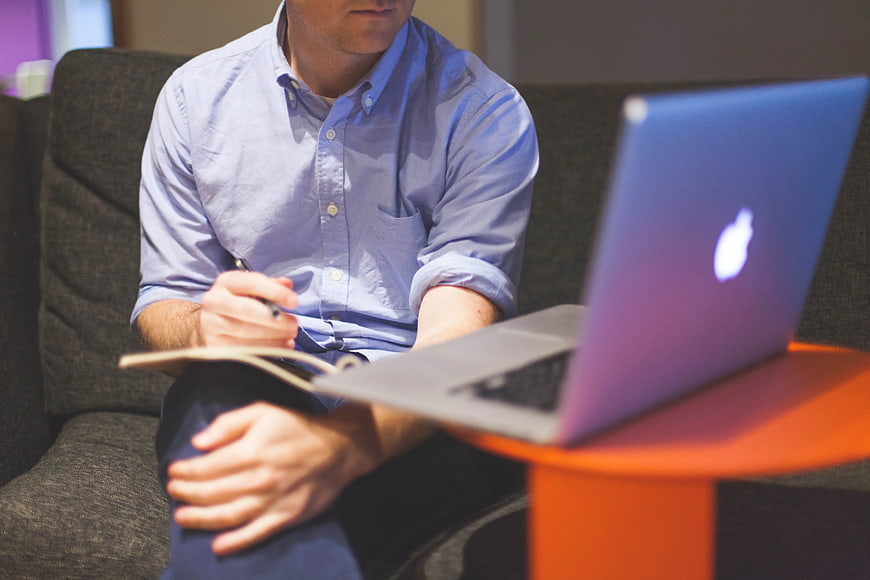 Man Writing in Notebook with Laptop on Couch