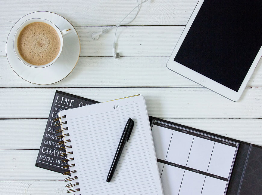 Flat lay of coffee, notebook, pen, and tablet on white wood