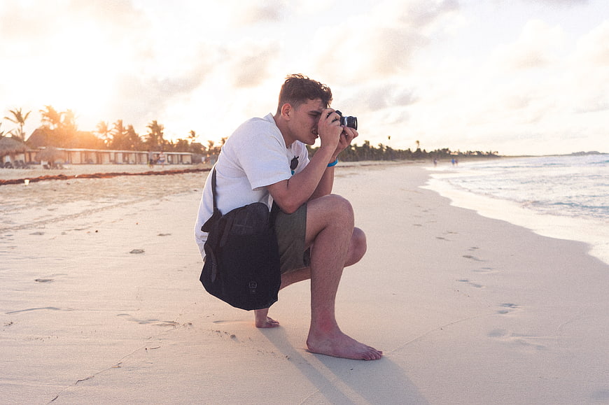 Young male photographer crouching on beach taking photo at sunset