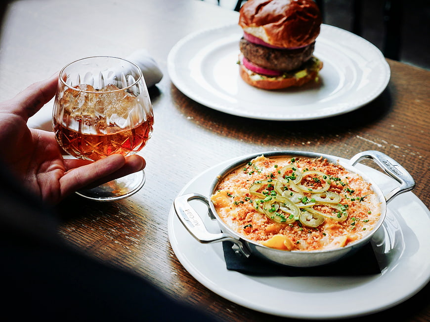 Hand holding cocktail with burger and mac and cheese on wooden table