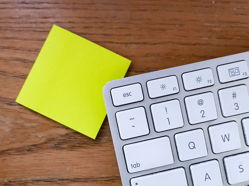 Yellow sticky note on wooden desk next to white keyboard