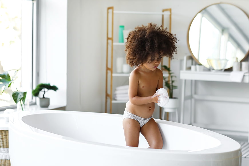 Cute child with curly hair playing with soap in bathtub