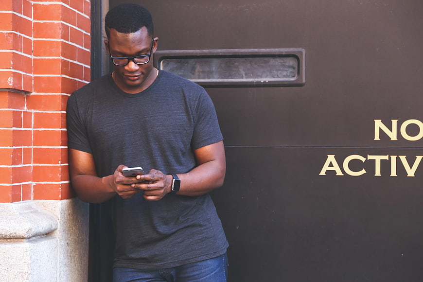 Man in Grey T-Shirt Using Smartphone Near Brick Wall