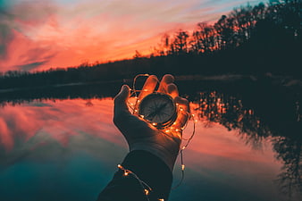 Hand holding compass with fairy lights at sunset lake