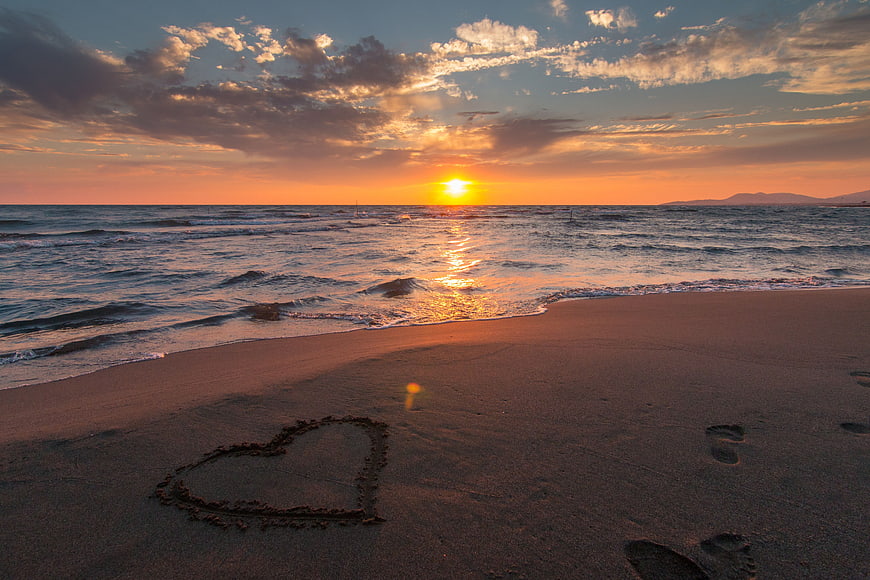 Heart drawn in sand on beach at sunset with footprints