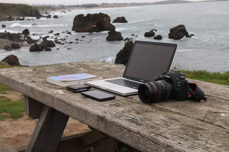 Photography gear and laptop on wooden table overlooking rocky ocean coast