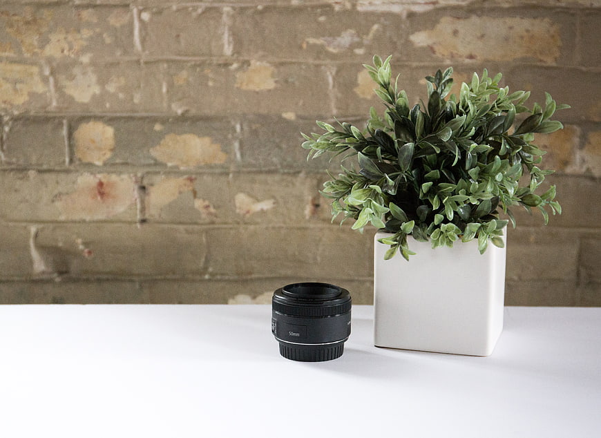 Camera lens and potted plant on white desk against brick wall
