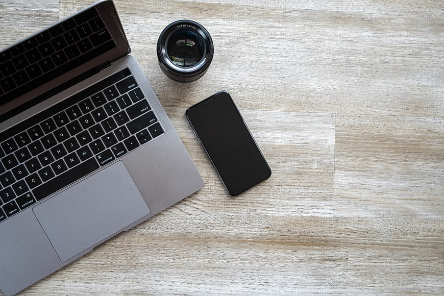 Flat lay of laptop, smartphone, and camera lens on wooden desk