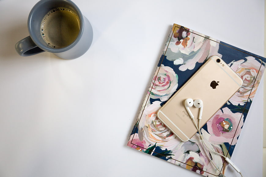 Flat lay of coffee mug, smartphone, and floral notebook on white desk