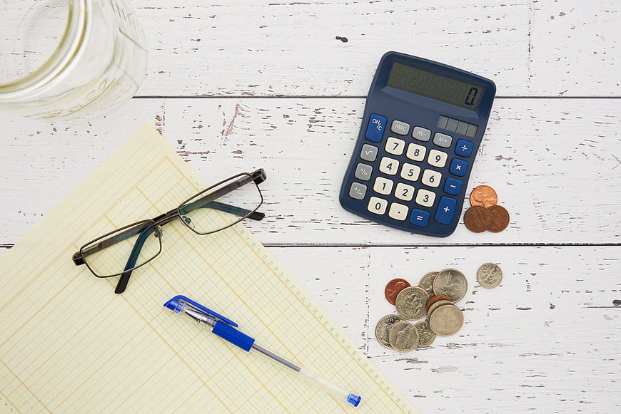 Top view of calculator, coins, glasses, and notebook on white wood