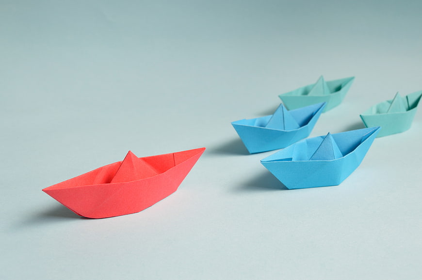 Red Origami Boat Leading Blue Paper Ships on Blue Background