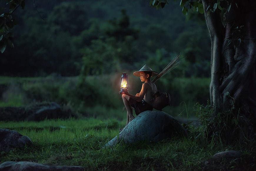 Young boy with lantern sitting on rock in forest at dusk