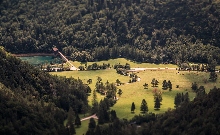 Aerial tilt-shift view of a lush green valley with a turquoise lake and dam