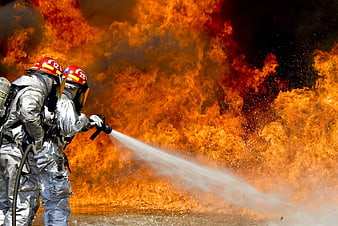 Two firefighters in silver suits extinguishing a massive fire