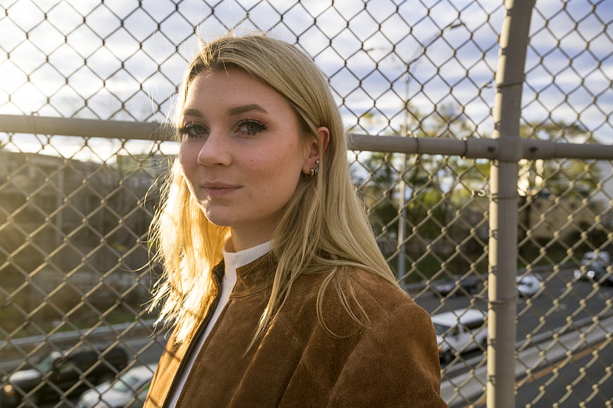 Blonde woman in brown jacket posing by chain-link fence