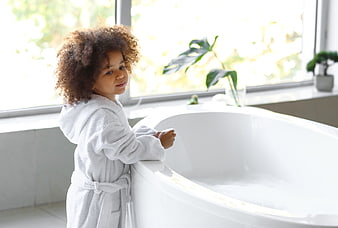 Cute child in white bathrobe standing by bathtub