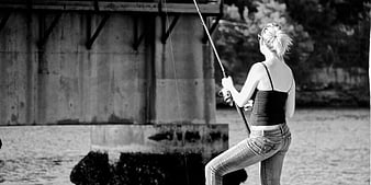 Woman fishing from riverbank under concrete bridge in black and white