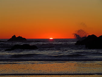 Dramatic Orange Sunset Over Ocean with Silhouetted Rocks and Splashing Waves