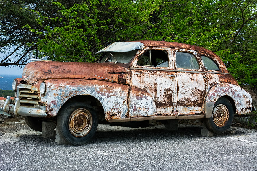 Rusty vintage car wreck on pavement with green trees