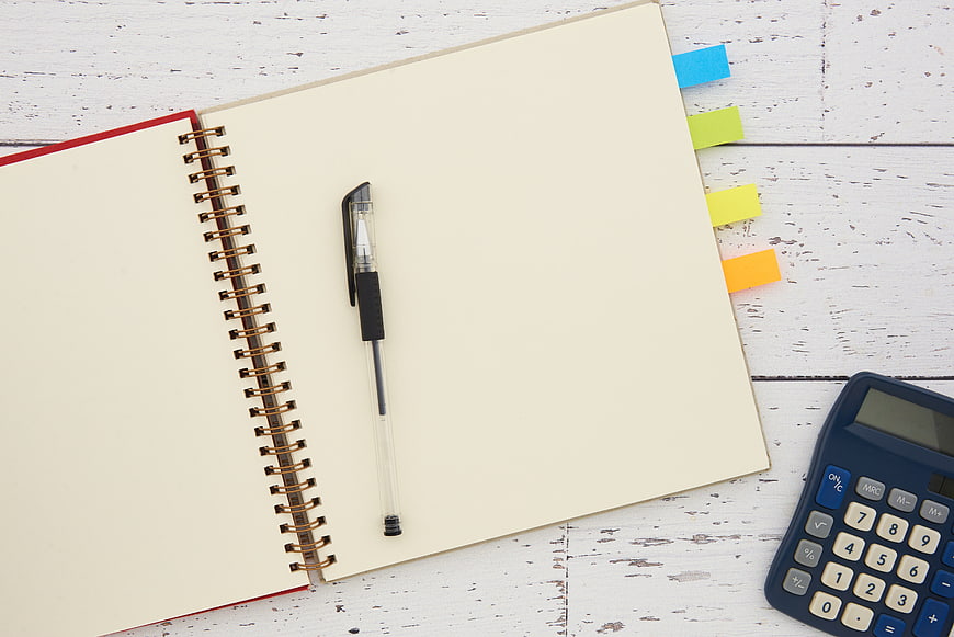 Flat lay of open notebook, pen, and calculator on white wood