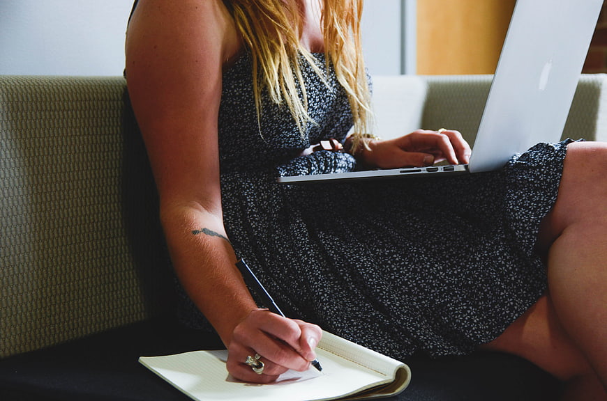 Woman Writing in Notebook with Laptop on Couch