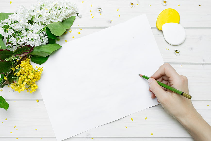 Hand holding green pencil over blank paper with spring flowers