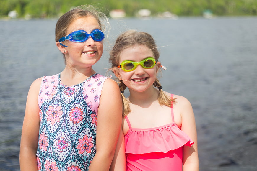 Two smiling girls in swimsuits and goggles by a lake