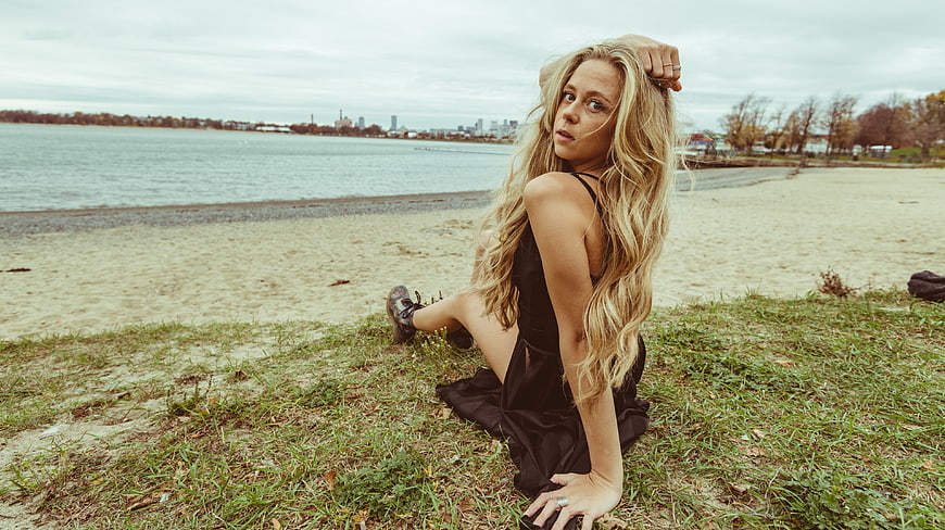 Blonde Woman in Black Dress Sitting on Beach with City Skyline