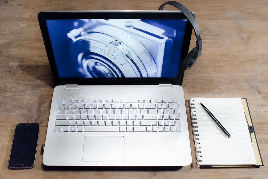 Laptop with camera screen, smartphone, notebook, and headphones on wooden desk
