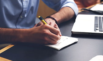 Close-up of hands writing notes on paper with laptop