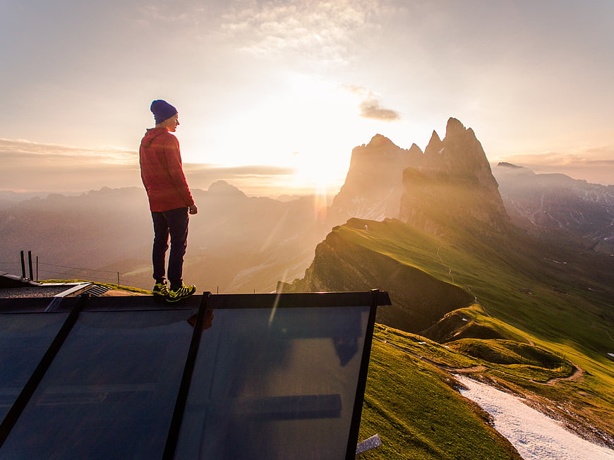Man standing on glass platform overlooking mountain peaks at sunset