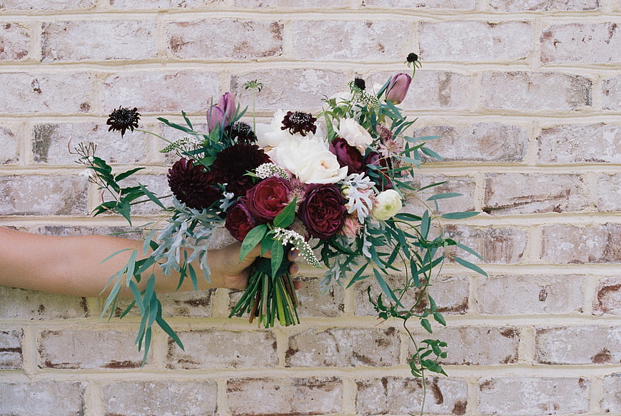 Hand Holding Dark Moody Wedding Bouquet Against Brick Wall