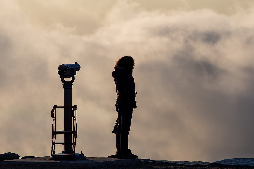 Silhouette of woman standing by telescope overlooking clouds