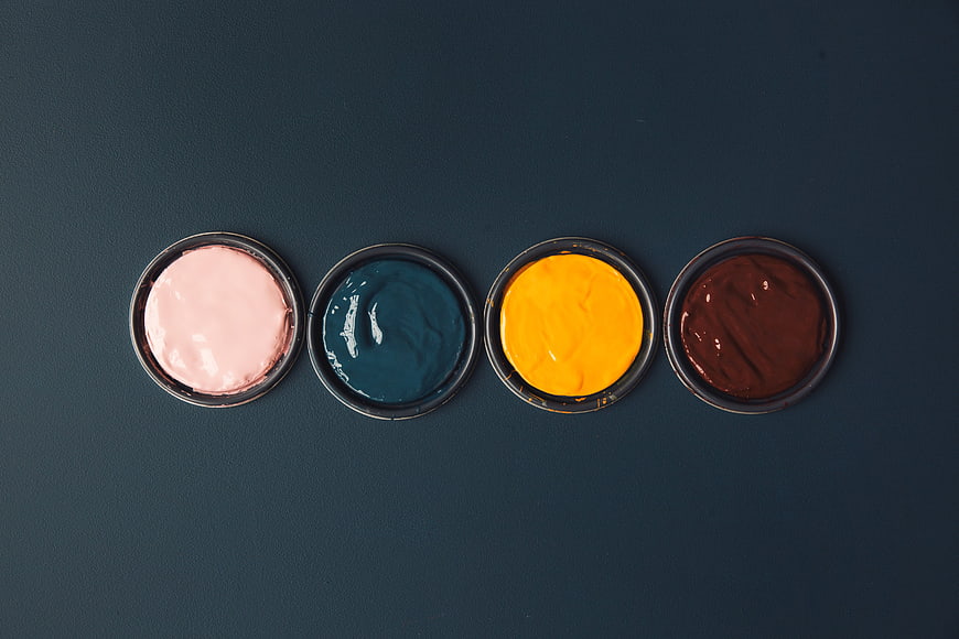 Top view of four open paint cans with pink, blue, yellow, and brown paint on dark background