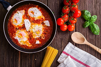 Shakshuka with eggs in tomato sauce, fresh basil, tomatoes, and pasta on wooden table