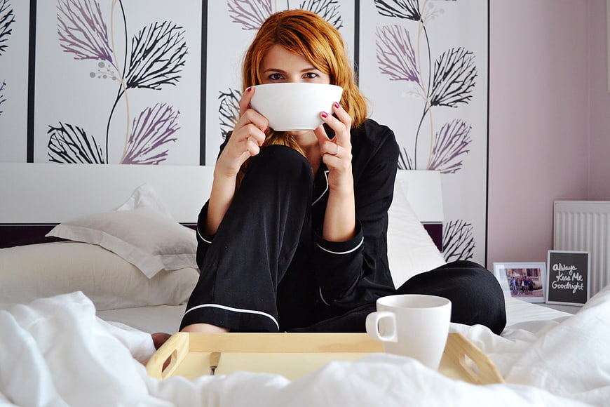 Woman in black pajamas eating breakfast in bed with floral wallpaper