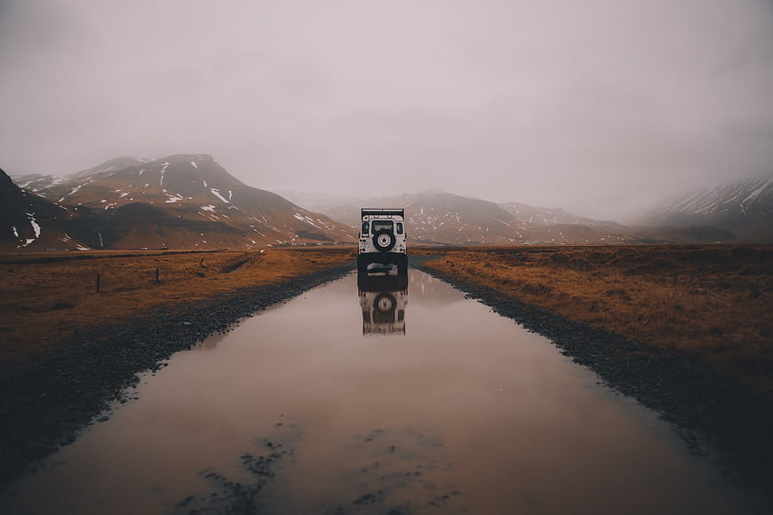 White Jeep Driving on Wet Road with Mountain Reflection
